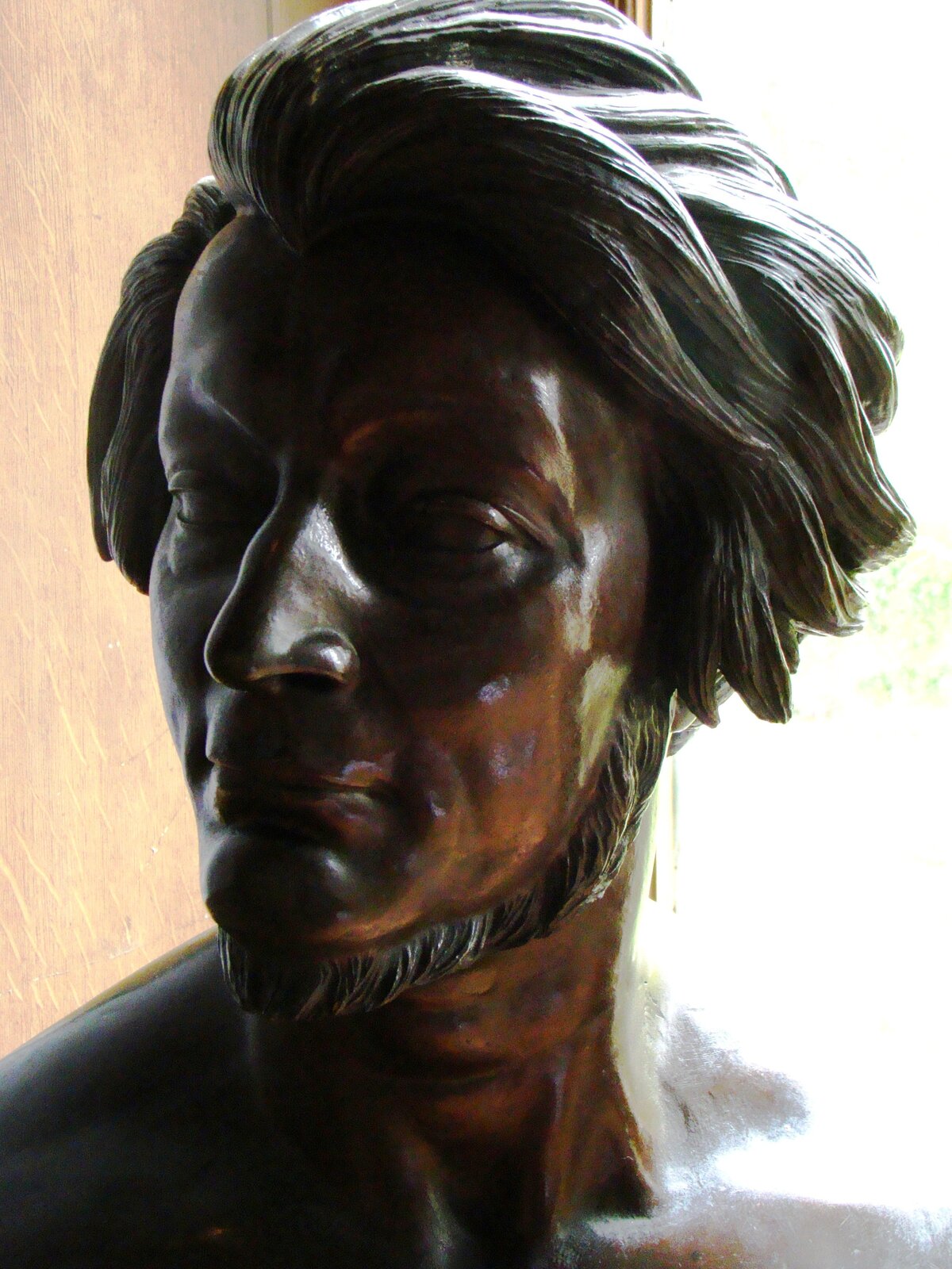 Zdjęcie przedstawia rzeźbę. To popiersie młodego mężczyzny z bujnymi, półdługimi włosami. Twarz mężczyzny jest pociągła, nos, wąski, wydatny. Mężczyzna ma krótki zarost poniżej podbródka.