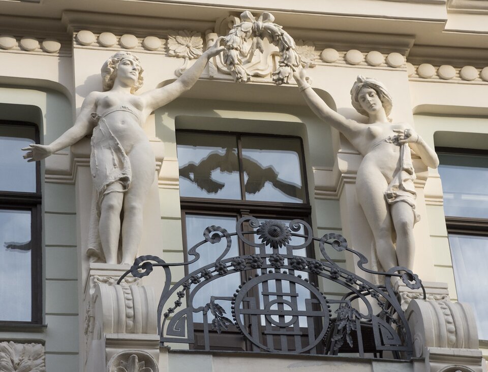 Ilustracja przedstawia fragment budynku z oknem i balkonem. Po dwóch stronach okna umieszczono rzeźby kobiet, unoszące  wspólnie wieniec nad oknem. Nad nimi biegnie dekoracyjny gzyms w koliste ornamenty. Balkon ma kutą, ozdobną barierkę. 