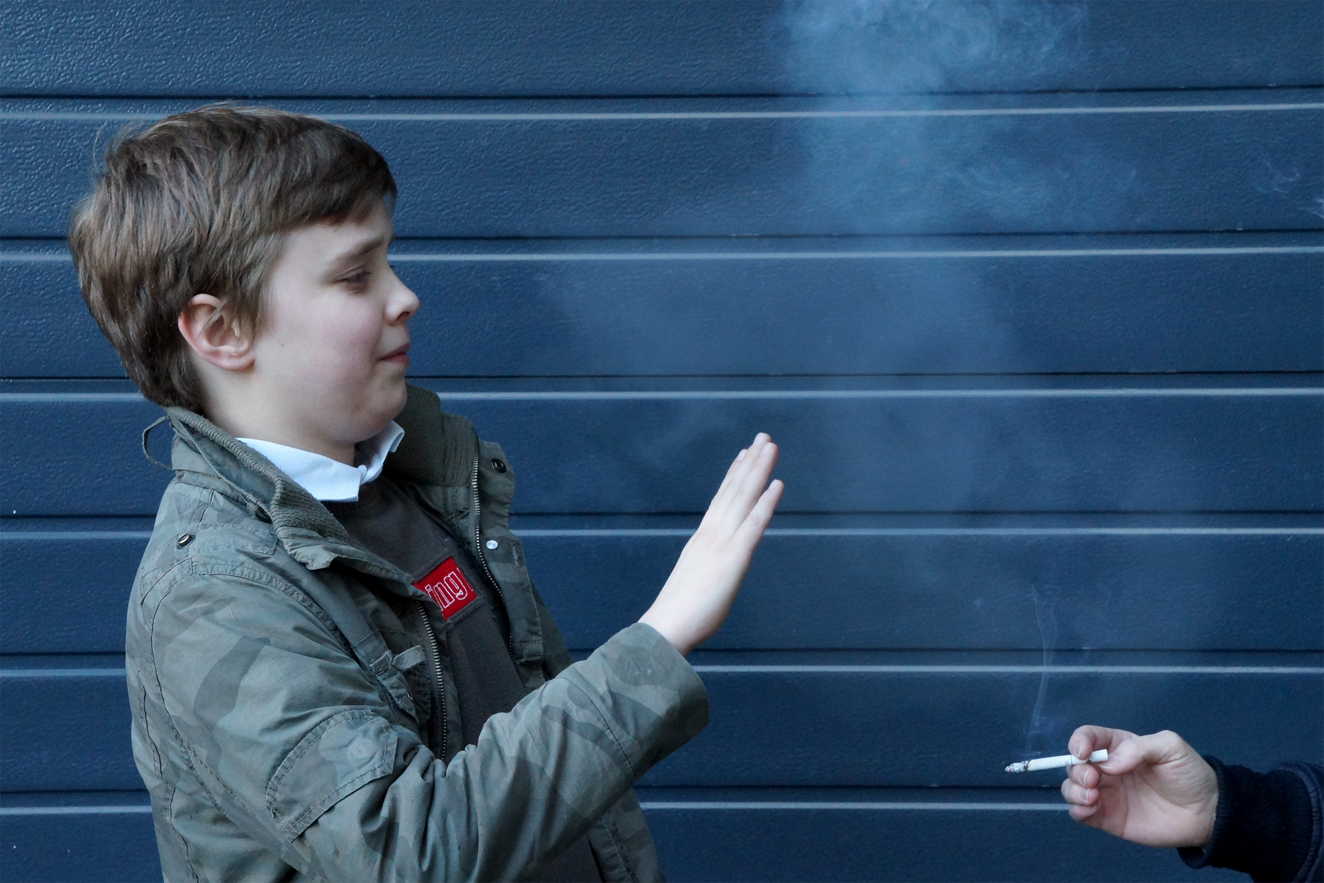 Fotografia przedstawia postać młodego chłopca z uniesioną dłonią, osłaniającego się od dymu papierosowego. W prawy dolnym rogu znajduje się dłoń innej osoby z zapalonym papierosem.