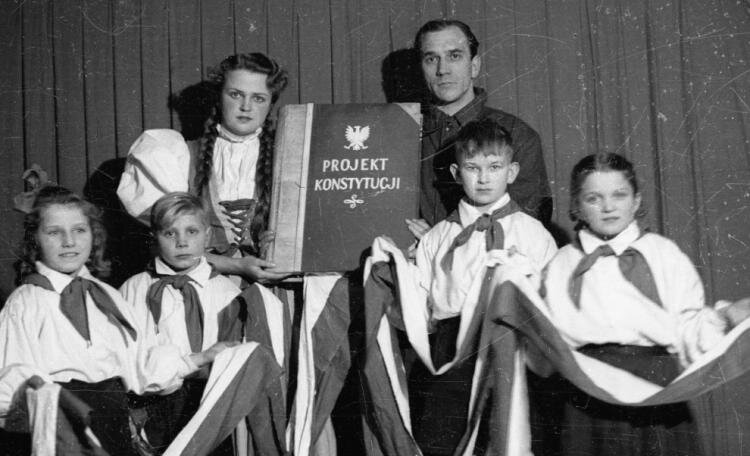Czarno‑białe zdjęcie przedstawia czworo dzieci ubranych w galowe stroje. Trzymają one długą biało‑czerwoną szarfę. Za nimi stoi kobieta i mężczyzna. Trzymają oni ogromną księgę z napisem projekt konstytucji.  Nad napisem znajduje się biały orzełek. 