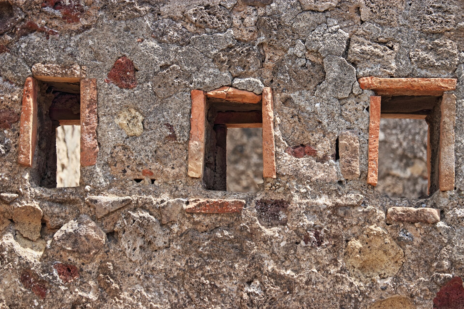 Fotografia przedstawia fragment ściany budynku, w której wykuto małe otwory okienne. Ściana budynku wykonana jest z różnokolorowych kamieni o chropowatej strukturze.