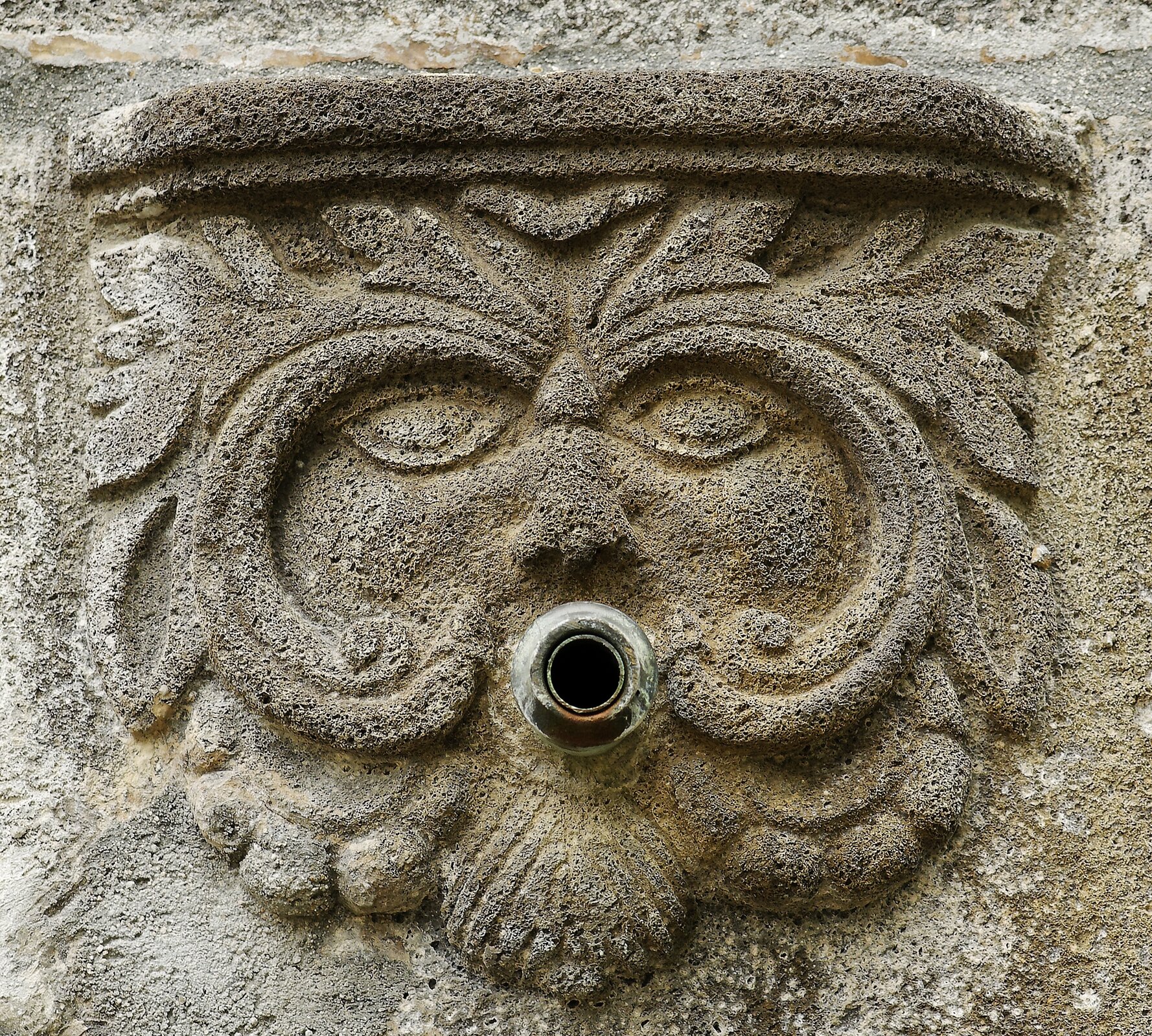 Zdjęcie przedstawia fragment fontanny w formie płaskorzeźby, wykonanej z jasnożółtego kamienia. Płaskorzeźba przypomina twarz. W miejscu ust jest otwór, z którego może wylewać się woda.