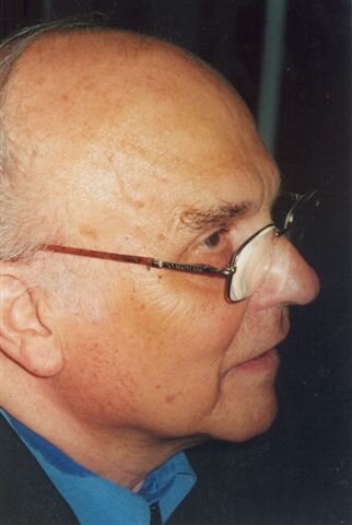Zdjęcie przedstawia prawy profil starszego mężczyzny w garniturze. Mężczyzna ma pełną twarz, wysokie czoło. Mężczyzna jest łysiejący, szpakowaty. Nosi okulary.