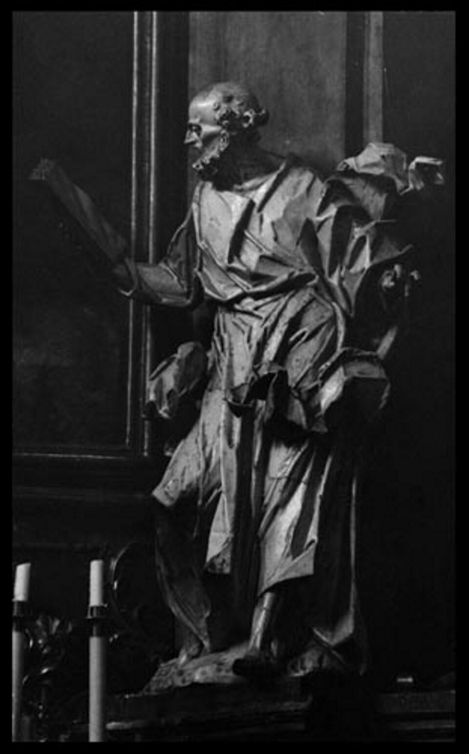 Ilustracja przedstawia posąg świętego Piotra na ołtarzu Przemieniania Pańskiego. Ilustracja ukazuje postać starszego mężczyzny. Postać stoi z głową zwróconą w prawą stronę w kierunku przedmiotu, który trzyma w prawej ręce. Lewa ręka postaci jest lekko uniesiona do góry.