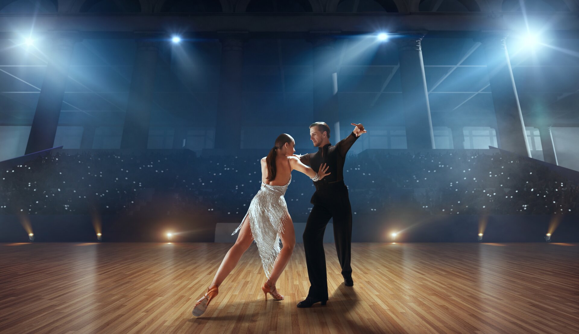 Fotografia przedstawia parę tańczącą Cha‑chę – latynoamerykański taniec, na konkursie tańca. Na pierwszym planie widoczne są dwie osoby – mężczyzna i kobieta. Mężczyzna ubrany jest w ciemną bluzkę z długim rękawem oraz golfem, a także czarne spodnie. Ma krótkie, zaczesane do tyłu włosy oraz krótką brodę. Ręce ma rozłożone na boki na wysokości ramion. Patrzy w stronę twarzy stojącej naprzeciwko niego kobiety, której długie, ciemne włosy spięte są w niski kucyk. Jej lewa ręka spoczywa na ręce mężczyzny, a prawa dotyka jego torsu. Tancerka stoi w rozkroku. Jej lewa noga jest cofnięta, opiera się na palcach, prawa jest zgięta w kolanie. Kobieta ma na sobie srebrną suknię odsłaniającą plecy oraz pomarańczowe buty na obcasie o średniej grubości. W tle widoczne są trybuny oraz lampy rozświetlające drewniany parkiet. 