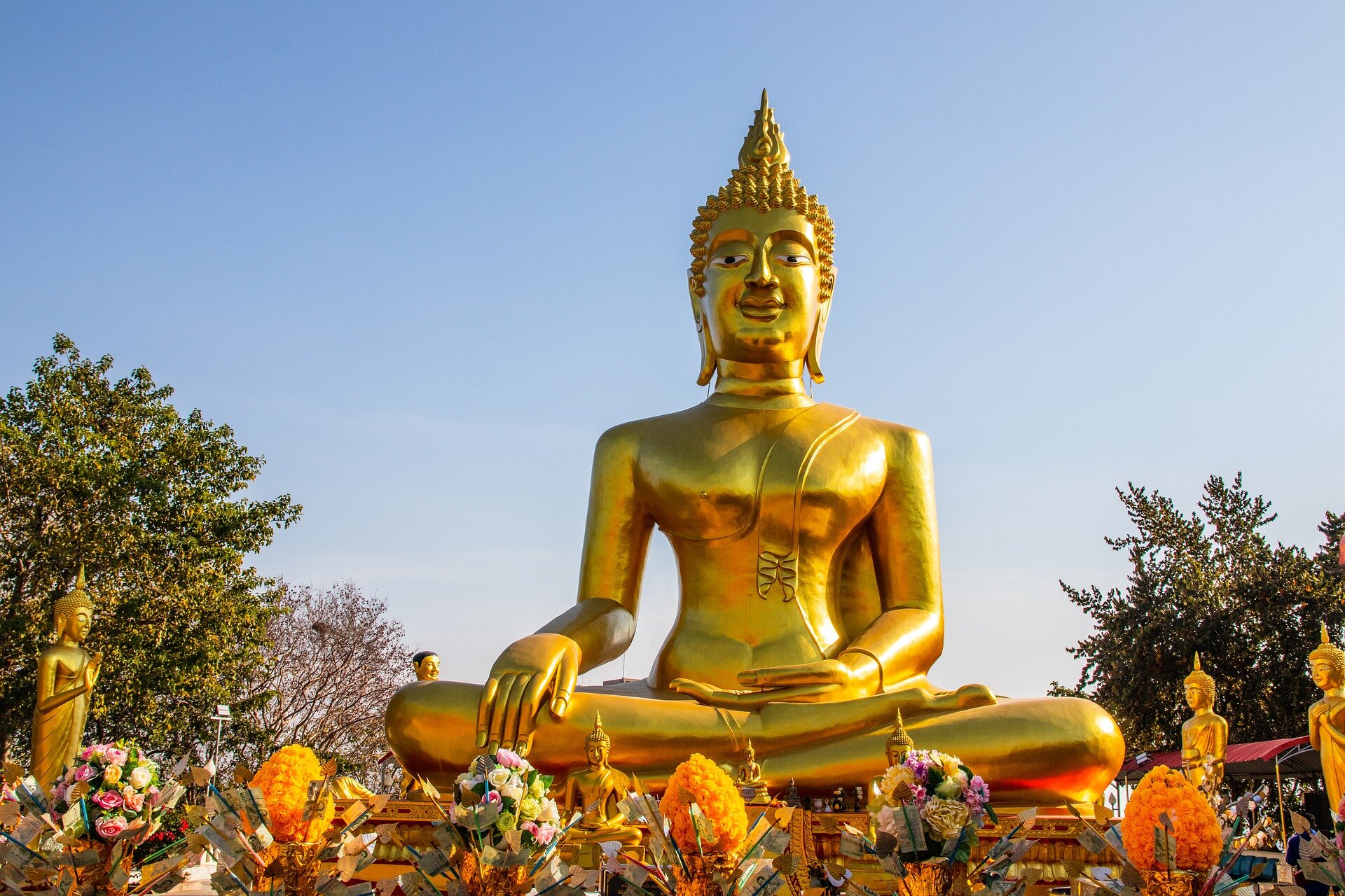 Zdjęcie przedstawia złoty, duży posąg Buddy. Budda siedzi w pozycji kwiatu lotosu, ma wysoko upięte włosy. Prawą dłoń opiera na prawym kolanie, lewą trzyma otwartą ku górze. Posąg jest złoty, znajduje się w plenerze. 