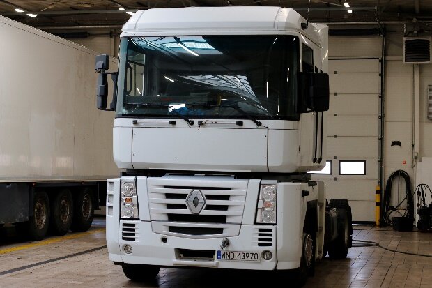 Ilustracja przedstawia ciągnik siodłowy w garażu. Ciągnik siodłowy jest koloru białego, obok ciągnika po lewej stronie widać naczepę także koloru białego.