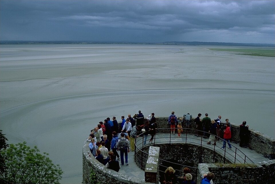Zdjęcie przedstawia wybrzeże wattowe we Francji podczas odpływu. Widoczne jest morze oraz strugi wody odpływające od lądu ku morzu . Na brzegu znajduje się okrągła wieża widokowa, na której szczycie ludzie obserwują odpływ.