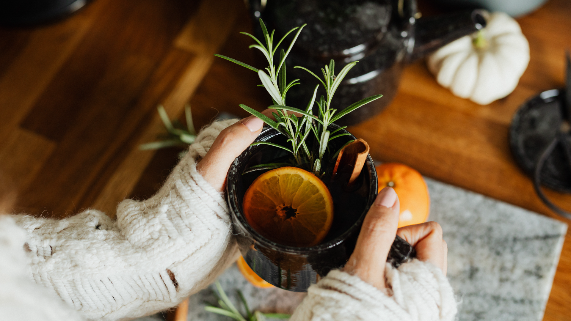 Zdjęcie przedstawia kubek z herbatą w kobiecych dłoniach. Zdjęcie wykonane jest z góry. W kubku widoczne są: plasterek cytryny, gałązka rozmarynu oraz laska cynamonu.