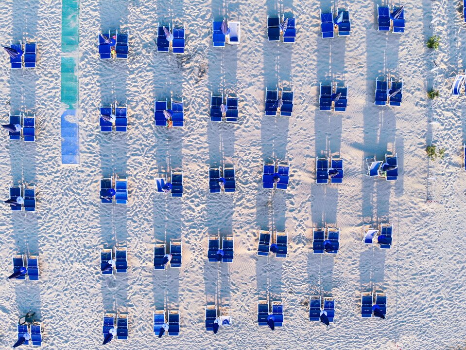 Zdjęcie przedstawia widok z góry na równo rozmieszczone niebieskie leżaki i parasole na jasnym piasku plaży.