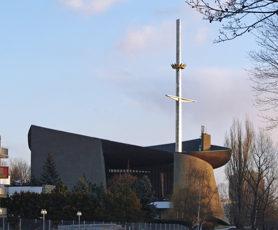Zdjęcie przedstawia modernistyczny budynek kościoła. Jest on owalny, z nierównym dachem. Na przedzie znajduje się wysoki krzyż. Budynek jest otoczony drzewami.