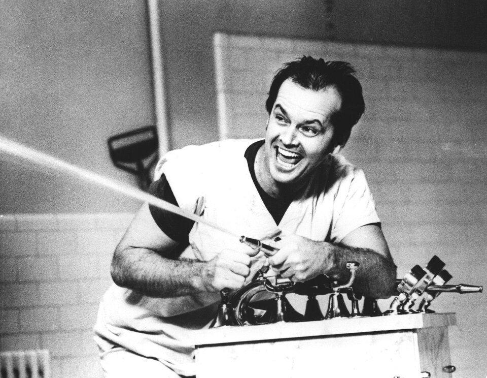 Zdjęcie z kadru filmu Lot nad kukułczym gniazdem. Widać na nim mężczyznę w średnim wieku, w szpitalnej pidżamie założonej na podkoszulek. Mężczyzna jest w łazience, trzyma w ręku wąż, z którego leci intensywny strumień wody. Mężczyzna cieszy się.