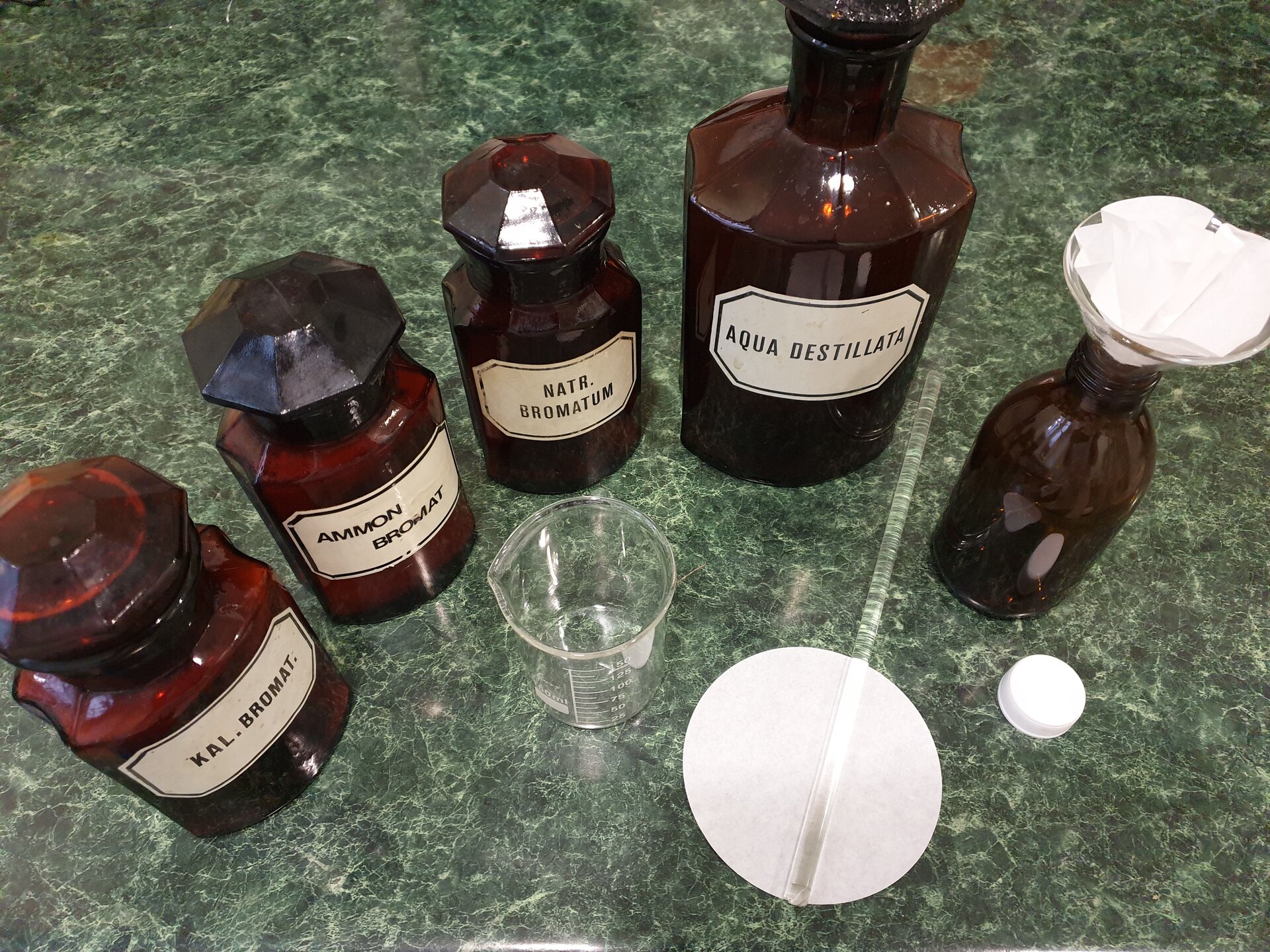 Rycina 3 przedstawia mieszanki. Na zdjęciu widoczne są stojące obok siebie na blacie szklane butelki ze szlifem, czyli ze szklanym dopasowanym zamknięciem w formie stożkowatej. Butelki mają różne rozmiary. Każda posiada podpisaną etykietę, między innymi: aqua destilata, czyli woda destylowana, kal. bromat., czyli bromek wapnia,  ammon bromat, czyli bromek amonu, natr bromatum, czyli bromek sodu. Obok stoi niewielka butelka, w którą włożono lejek z sączkiem. Obok znajduje się niewielka zlewka, sączek oraz szklane mieszadełko w postaci patyczka.