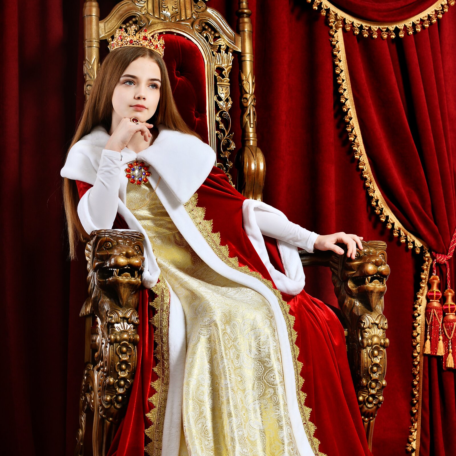 Zdjęcie przedstawiające młodą dziewczynę siedzącą na tronie. Ma ona długie, brązowe włosy, ubrana jest w czerwoną szatę i złotą sukienkę. Na głowie ma złotą koronę.