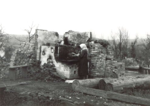 Zdjęcie przedstawia kobietę, która stoi przy kamiennym piecu. Piec otaczają zgliszcza domu, z którego pozostał jedynie fragment ścian. Na ziemi leżą cegły oraz pale drewna. Kobieta ma prostą sukienkę i chustę na głowie.