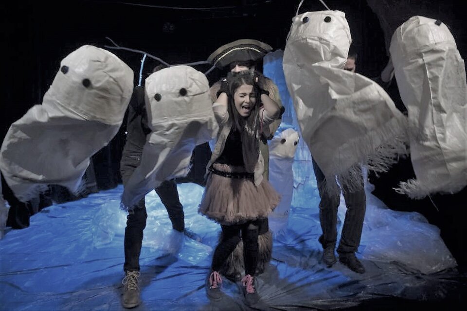 Zdjęcie przedstawia scenę teatralną wysłaną białą folią imitującą śnieg, na którym stoi dziewczyna otoczona przez bałwany. Dziewczyna boi się, zakrywa uszy i krzyczy. Jest ubrana współcześnie, a sceneria jest jak ze snu. Dwóch aktorów stojących za nią, animuje 4 duże lalki - bałwany, które otaczają dziewczynę od tyłu i po bokach. Są zrobione z postrzępionych worków, mają przyklejone czarne kółka - oczy. 