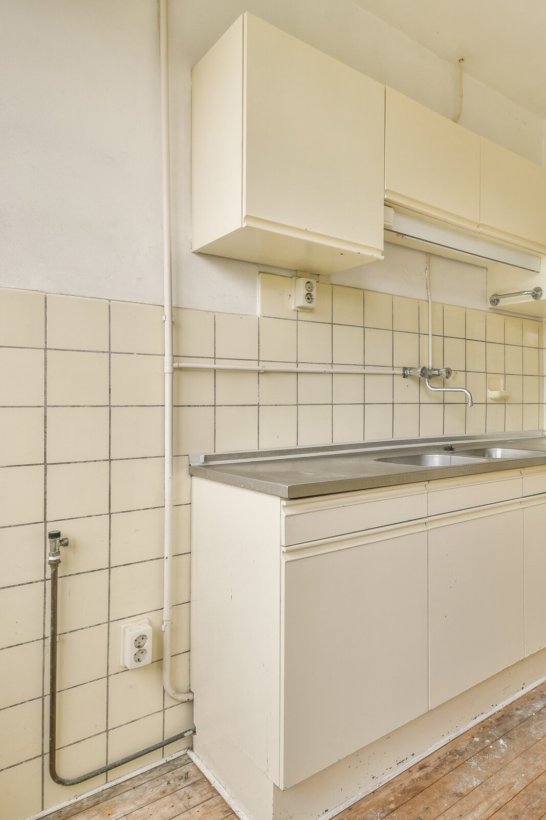 Na zdjęciu widoczne są przewody instalacji wodnej rozmieszczone na ścianie. Od pionowej rury odchodzi pozioma zakończona baterią umieszczoną nad zlewem.