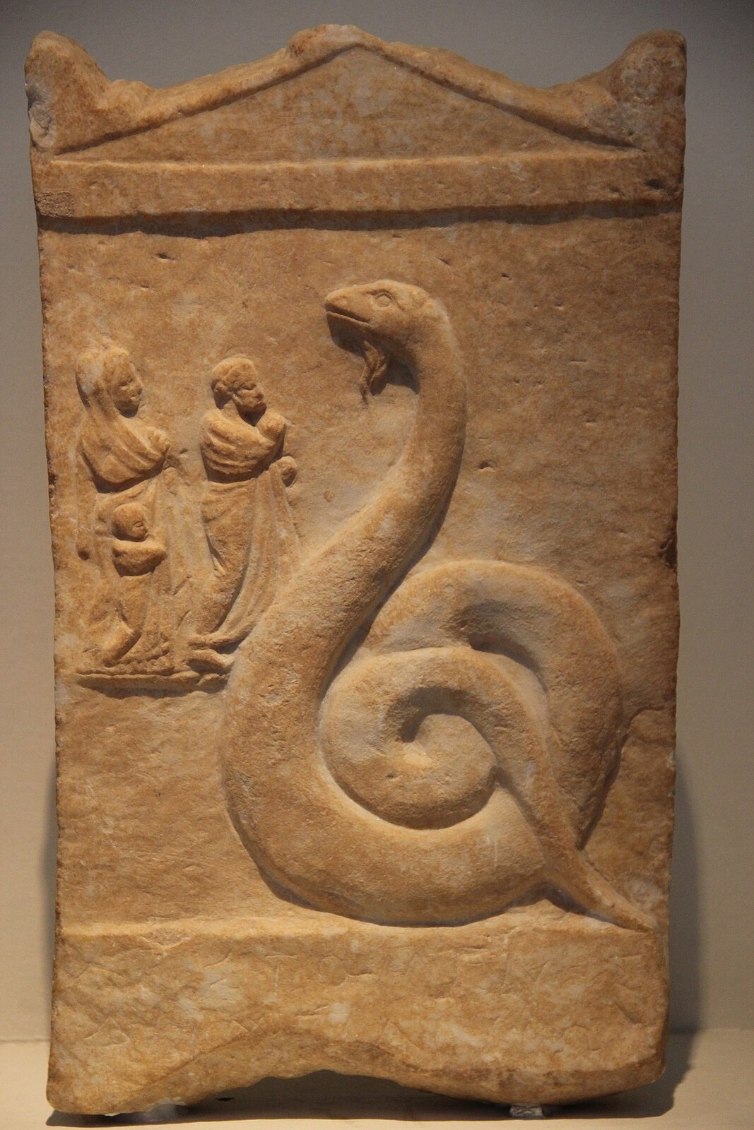 Fotografia przedstawia kamienną płytkę o kształcie prostokąta, którego górna krawędź jest wycięta w trójkątny wzór. Na ściance płytki wyrzeźbiony jest duży wąż pożerający człowieka. W tle dwie osoby dorosłe i dziecko przyglądają się scenie.