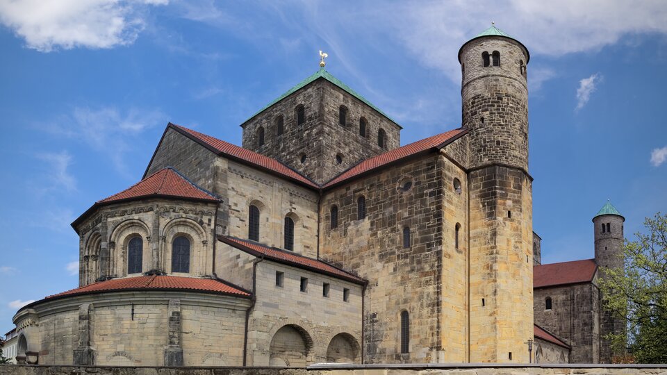 Ilustracja przedstawia kościół św. Michała w Hildesheim. Zdjęcie ukazuje budynek składający się z kilku brył o różnej wysokości, z dwoma transeptami i wieżami w typie ołówkowym. Ba wszystkich ścianach świątyni znajdują się niewielkie otwory okienne zakończone półokrągłym łukiem. Na skrzyżowaniu naw znajduje się wieża na planie kwadratu. Dach korpusu budowli jest dwuspadowy. Zdjęcie jest wykonane od strony apsydy.