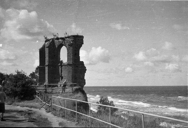 Archiwalne, czarno białe zdjęcie ruin kościoła w Trzęsaczu z 1971 r. Obraz przedstawia gruzy w kolejnym stadium degradacji ze względu na osuwające się urwisko. W murach otwór okienny bez szyb. Wzdłuż ścieżki prowadzących do ruin barierka nad brzegiem osuwiska. W tle morze. 