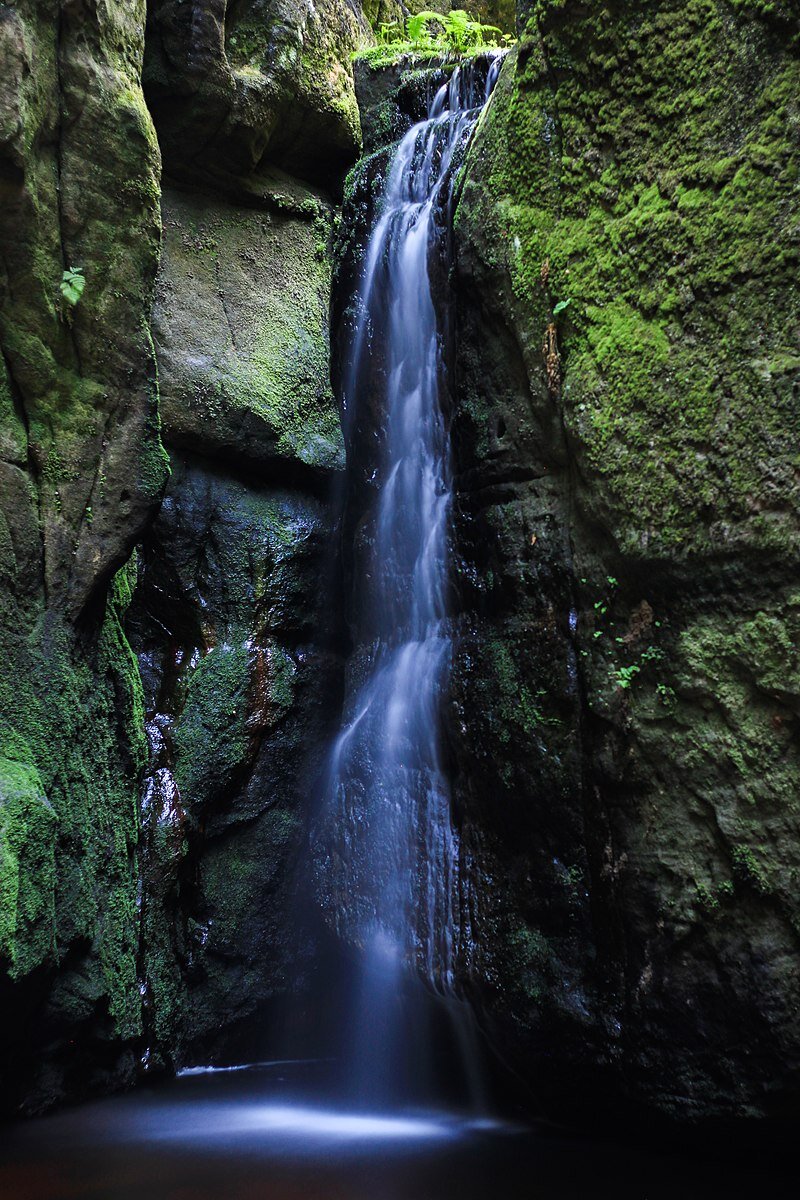 Fotografia wodospadu spływającego z pionowej skały. Po bokach skały pokryte porostami.