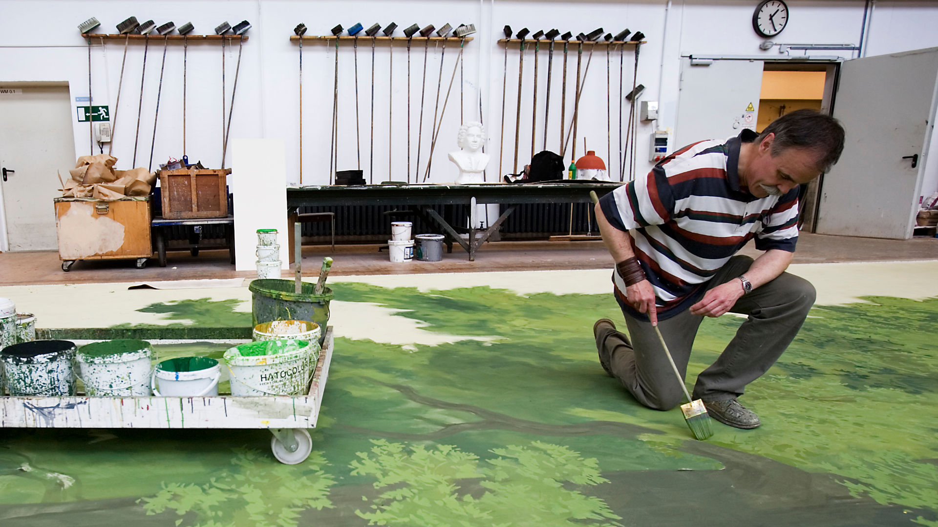 Zdjęcie przedstawia mężczyznę, który maluje pędzlem roślinne wzory na olbrzymim płótnie. Płótno leży na ziemi. Obok znajduje się wózek z farbami.