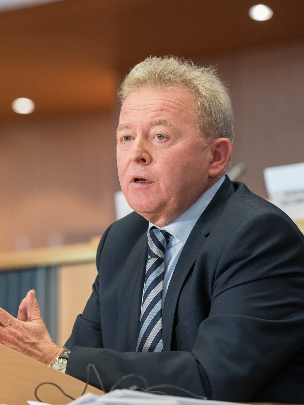 Fotografia przedstawiająca mężczyznę w sile wieku. Postać siedzi, ukazana jest od pasa w górę. Mężczyzna pokazuje lewy półprofil, ma okrągłą twarz, gęste włosy nieco dłuższe z przodu. Lekko marszczy czoło i unosi gęste brwi. Ma duże oczy i nos oraz otwarte usta. Mężczyzna jest ubrany w koszulę i marynarkę. Na szyi ma zawiązany krawat. Spod lewego mankietu wystaje zegarek z dużą tarczą. Ręce opiera o pulpit, dłonie ma złączone.