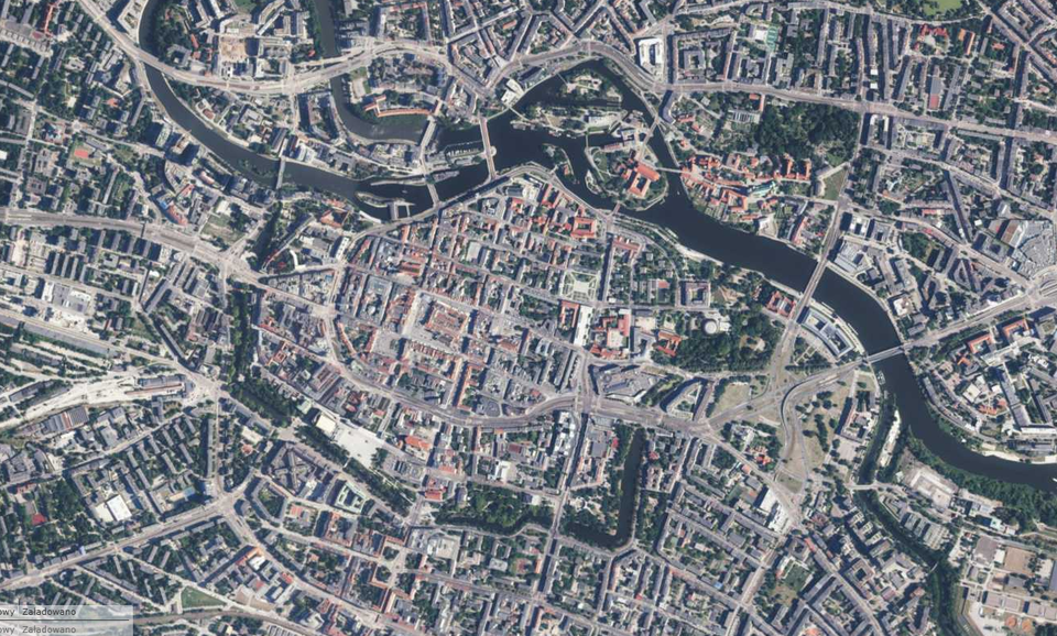 Fotografia przedstawia lotnicze zdjęcie centrum Wrocławia. Dobrze widoczny układ ulic i zabudowy. Widać czerwone i szare dachy. Z prawego dolnego rogu do lewego górnego rogu wije się rzeka Odra. Można dostrzec również mosty i drzewa.