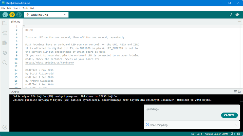 Zrzut ekranu przedstawia środowisko Arduino podczas wgrywania programu „Upload” do płytki Arduino Uno. W głównym oknie otwarty jest przykładowy szkic „Blink.ino” z widocznym komentarzem opisującym program. W dolnej części ekranu, w czarnym oknie „Output”, wyświetlana jest informacja o zużyciu pamięci (programowej: 924 bajty, dynamicznej: 9 bajtów) oraz statusie procesu. Po prawej u dołu pojawia się komunikat „Uploading...” z możliwością anulowania „Cancel”. Pasek statusu w prawym dolnym rogu pokazuje „Arduino Uno on COM1”.