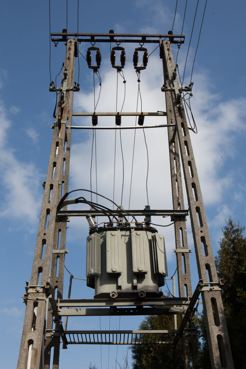 Fotografia przedstawia wysoki, betonowy słup z podłączonymi czarnymi kablami i popielatą metalową skrzynią na środku - jest to transformator.