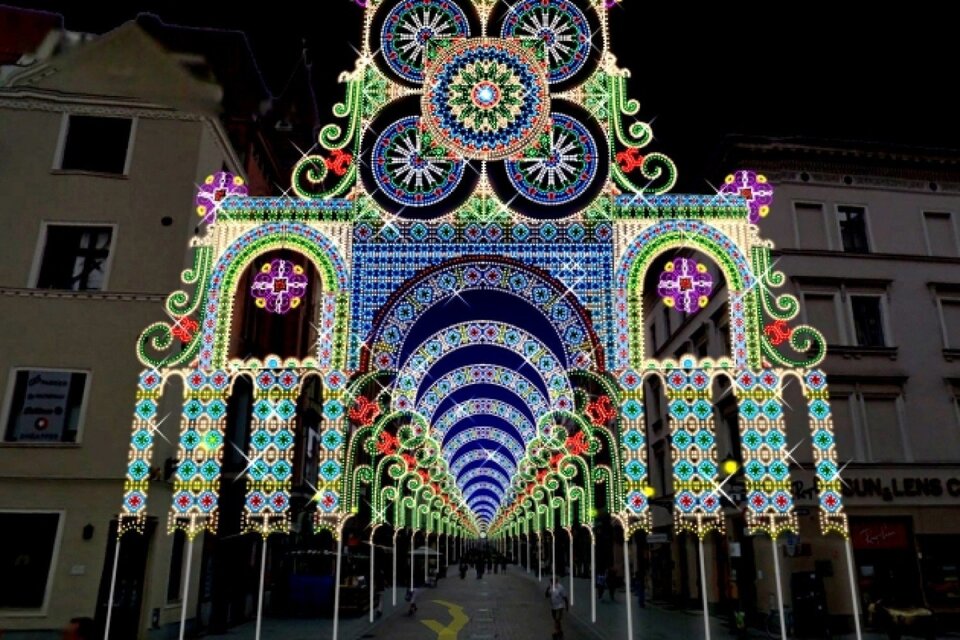 Ukazana zostaje fotografia przedstawiająca przejście pomiędzy dwoma beżowymi budynkami. Przejście oświetlone jest wieloma kolorowymi światłami. Światła składają się na spójną całość tworząc korytarz świetlny imitujący świetliste niebo.