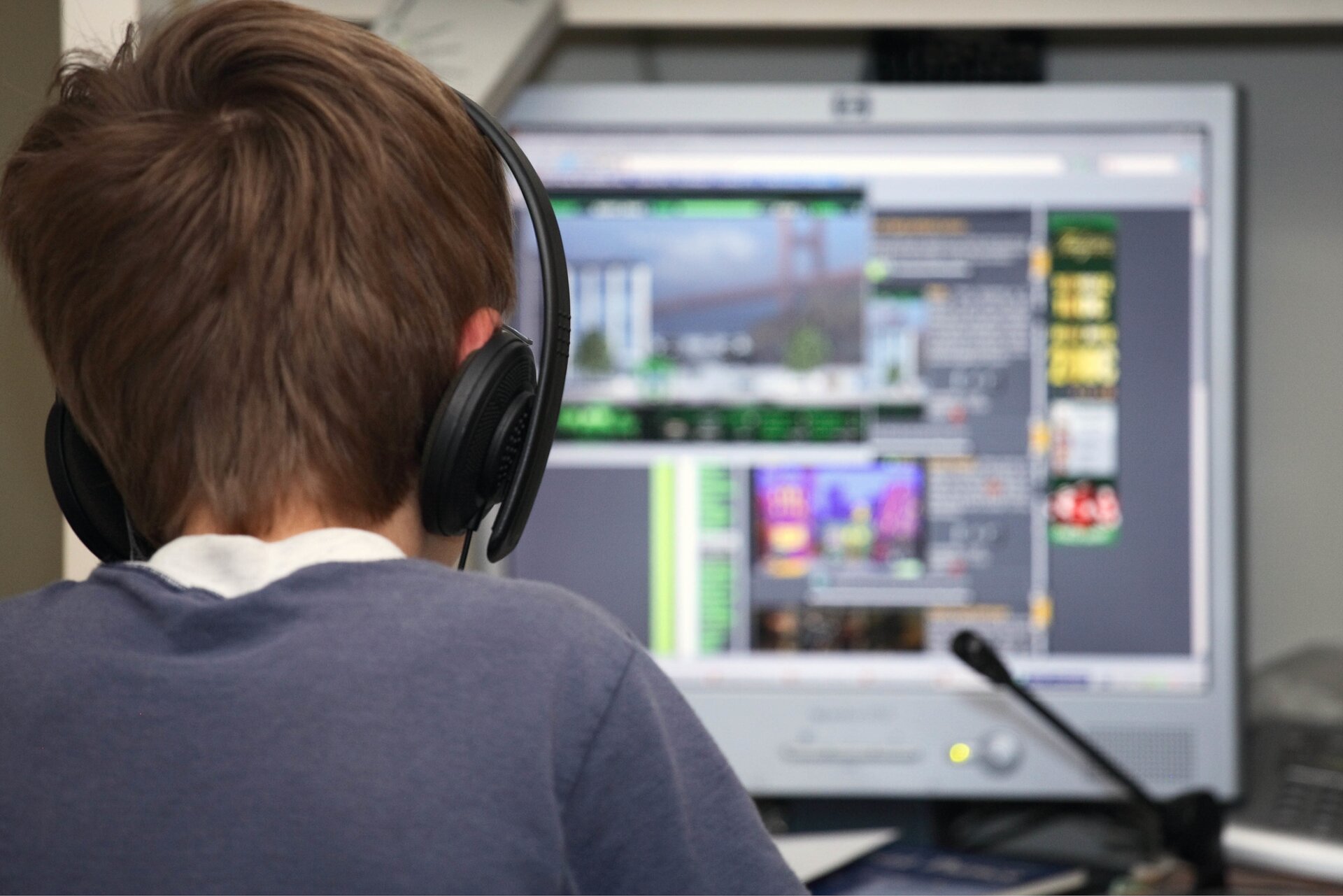 Zdjęcie ukazuje plecy chłopca siedzącego przed komputerem. Przed ekranem stoi mikrofon. Na ekranie widać trzy otwarte okna stron internetowych. Chłopiec ma na uszach założone duże słuchawki.