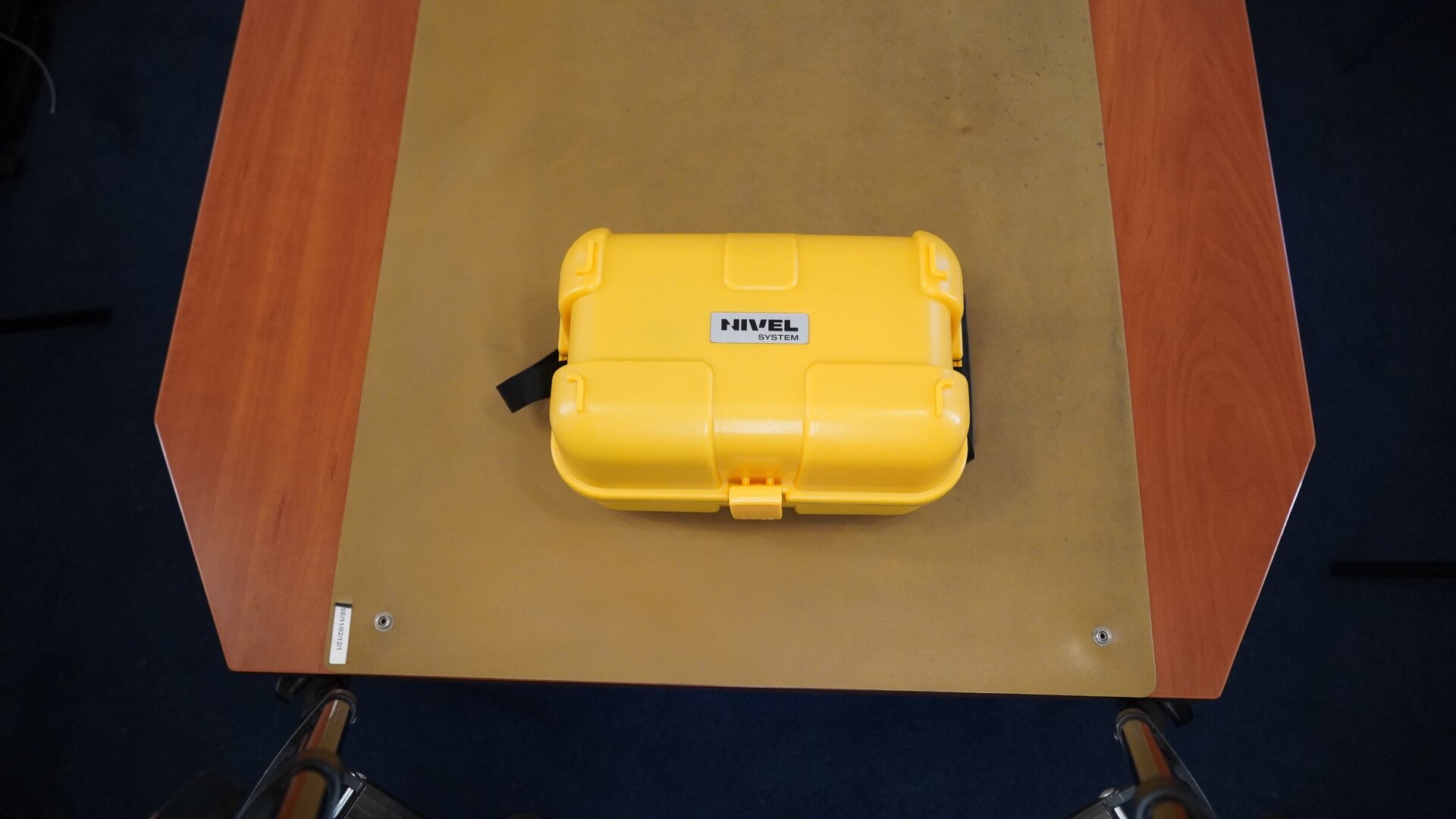 Zdjęcie przedstawia żółtą skrzynkę w kształcie walizki wykonaną z tworzywa sztucznego. Skrzynka została umieszczona na blacie. Na skrzynce widnieje plakietka z napisem: „Nivel system”.