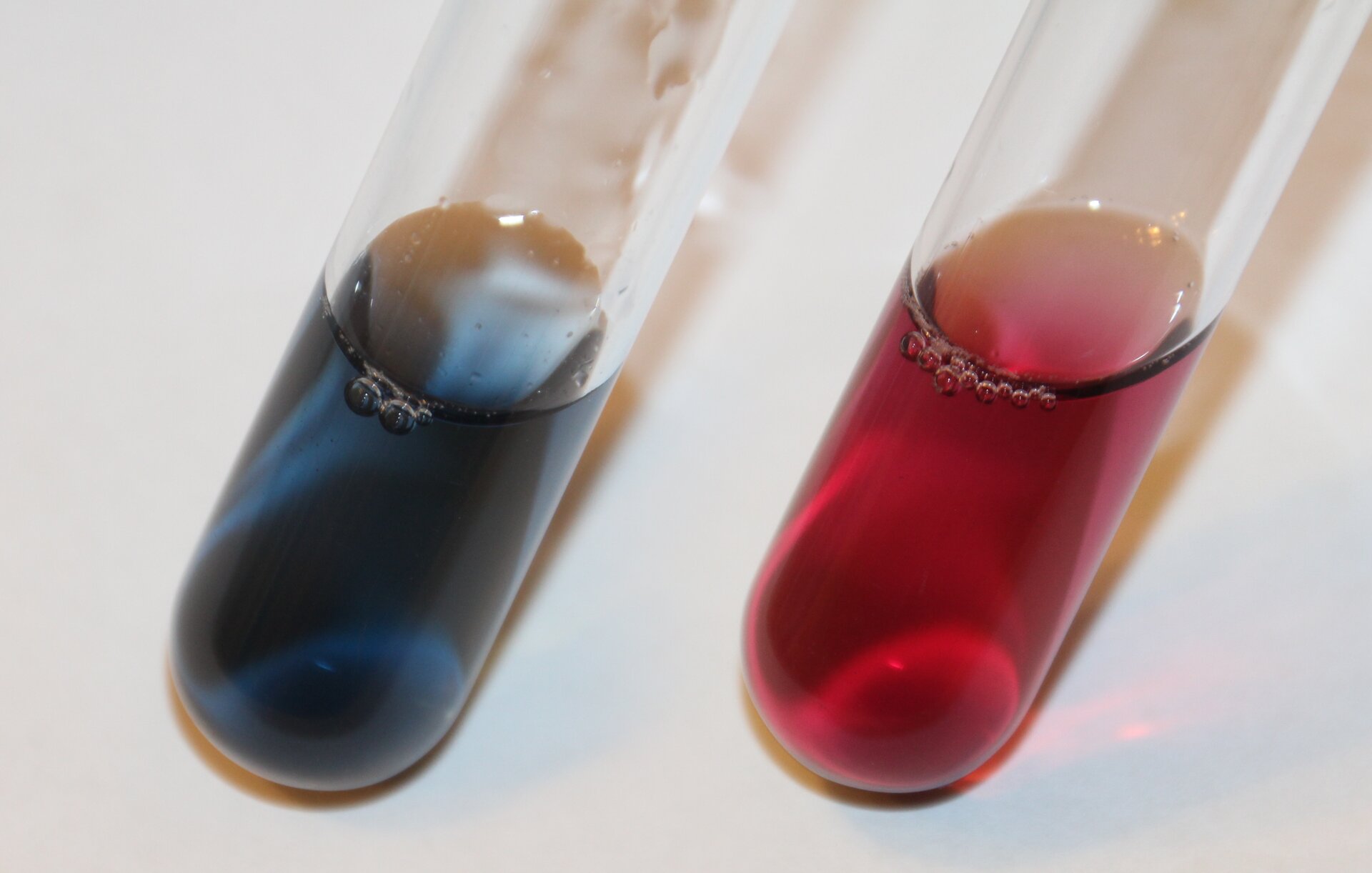 Na zdjęciu są dwie probówki. W jednej jest przezroczysty niebieski roztwór, w drugiej przezroczysty czerwony roztwór.  