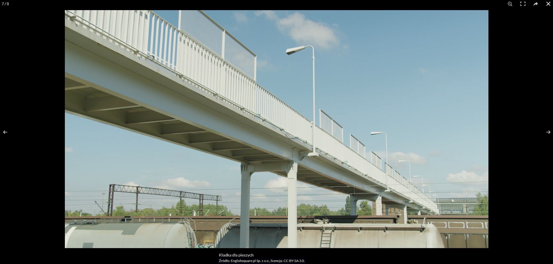 Zrzut ekranu przedstawia przykładowy widok zdjęcia w galerii. Zdjęcie jest widoczne w trybie podglądu. Na środku znajduje się zdjęcie kładki dla pieszych. Pod zdjęciem jest jego podpis oraz źródło. W górnym lewym rogu jest licznik zdjęcia, wskazujący, że to zdjęcie siódme z ośmiu. Przy prawej i lewej krawędzi znajdują się strzałki do przełączania się między zdjęciami. W górnym prawym rogu widoczne są cztery przyciski: zoom, przełączanie pełnego ekranu, udostępnianie oraz zamknięcie podglądu.