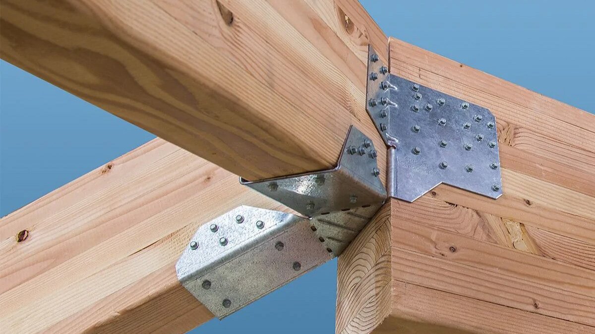 Zdjęcie przedstawia punkt styku trzech belek, które tworzą kąty ostre. Mocuje je metalowa blaszka z wypustkami, która znajduje się na wszystkich trzech belkach, przytwierdzona do nich od boku i od spodu.