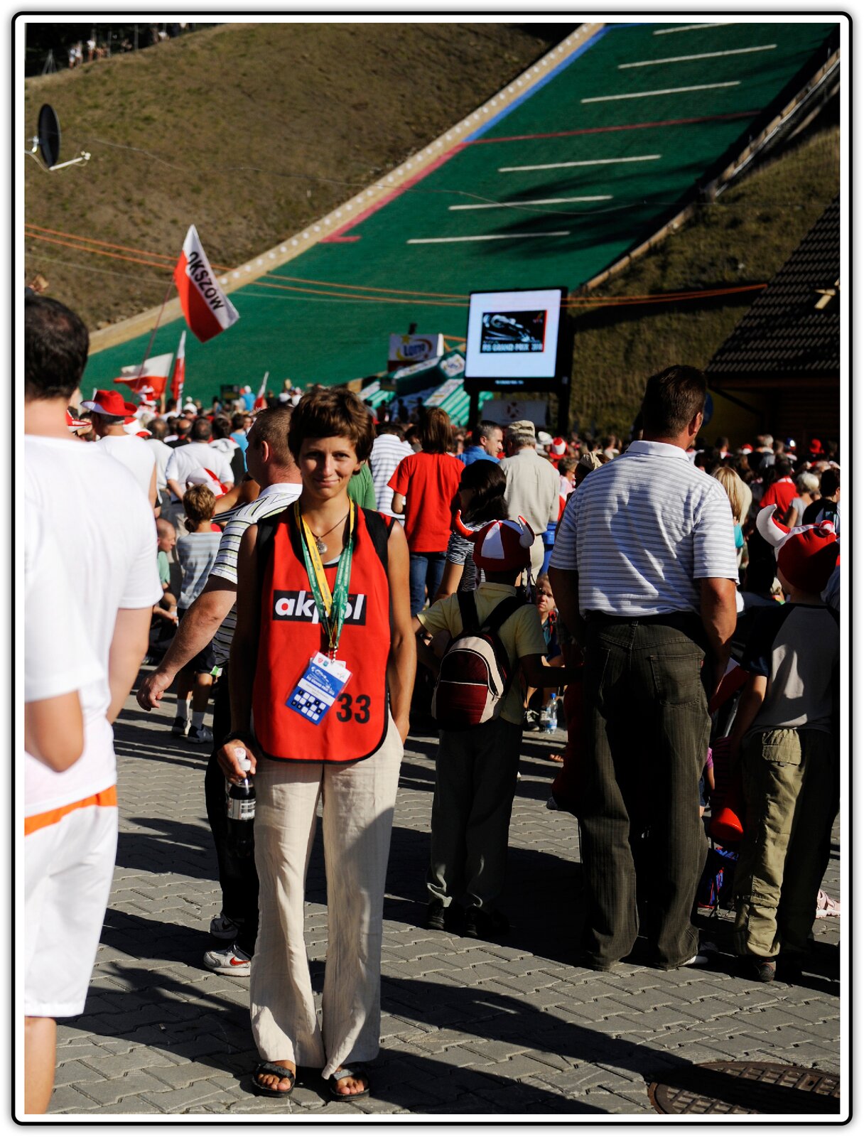 Fotografia przedstawia ludzi zgromadzonych przy torze skoczni, w tle ilustracji widać tor, który otaczają ludzie w barwnych strojach trzymający polskie flagi w dłoniach, nad głowami zebranych znajduje się telebim, na którego ekranie widać skaczącego zawodnika. Widzowie są zwróceni tyłem do oglądającego fotografię. Tylko jedna osoba wyróżnia się z tłumu.To młody mężczyzna odwrócony przodem. Jest uśmiechnięty, ubrany w czerwoną koszulkę bez rękawów i beżowe długie spodnie, na szyi ma zawieszone na grubych tasiemkach identyfikatory, a na nogach  sandały, jedną rękę trzyma w kieszeni, w drugiej natomiast ma butelkę z jakimś napojem. Wygląda na osobę zadowoloną i dobrze bawiącą się na imprezie sportowej.