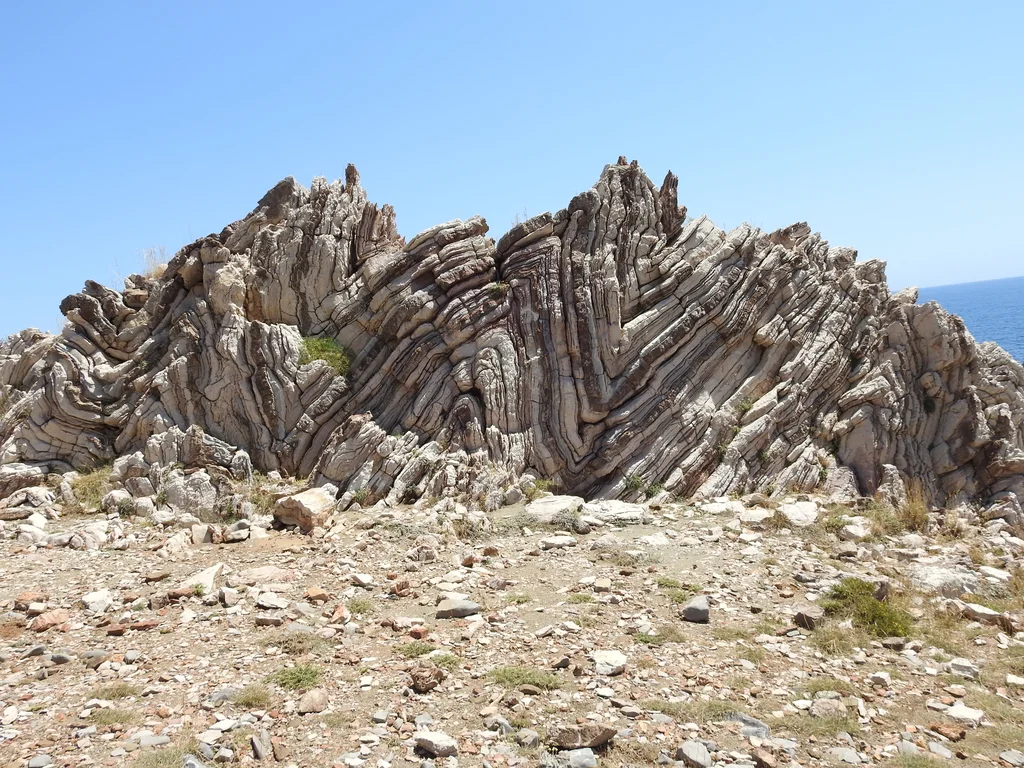 Zdjęcie przedstawia sfałdowaną formację skalną. Warstwy skalne układają się w nieregularne łuki. Przed formacją jest płaski ziemisto kamienisty teren z kępkami trawy.