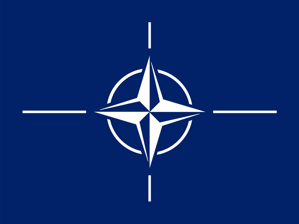 Grafika przedstawia flagę NATO. Na granatowym tle na środku znajduje się biały pierścień symbolizujący jedność, a w nim róża kompasowa, symbolizująca dążenie do zapewnienia bezpieczeństwa ze wszystkich stron.