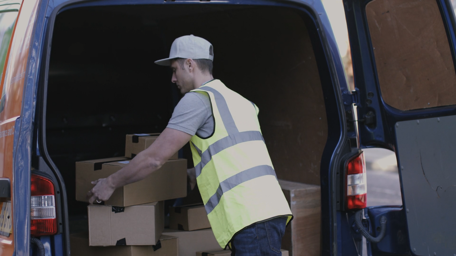 Zdjęcie przedstawia mężczyznę w białej czapce oraz kamizelce odblaskowej, który wykonuje załadunek samochodu dostawczego kartonowymi pudełkami.