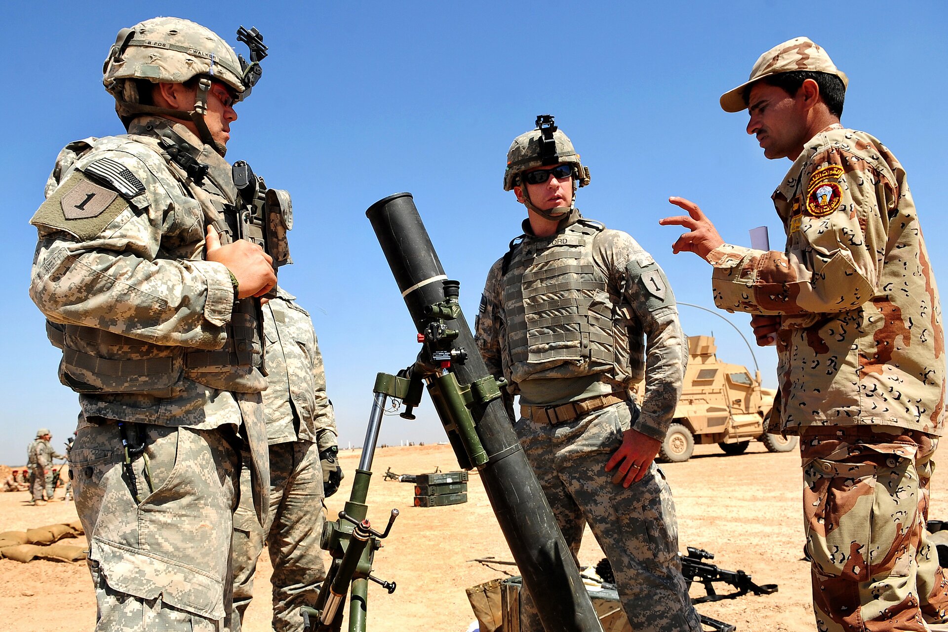 Zdjęcie przedstawia trzech rozmawiających ze sobą żołnierzy. Za nimi pojazd wojskowy, broń i pozostali żołnierze. Krajobraz pustynny.