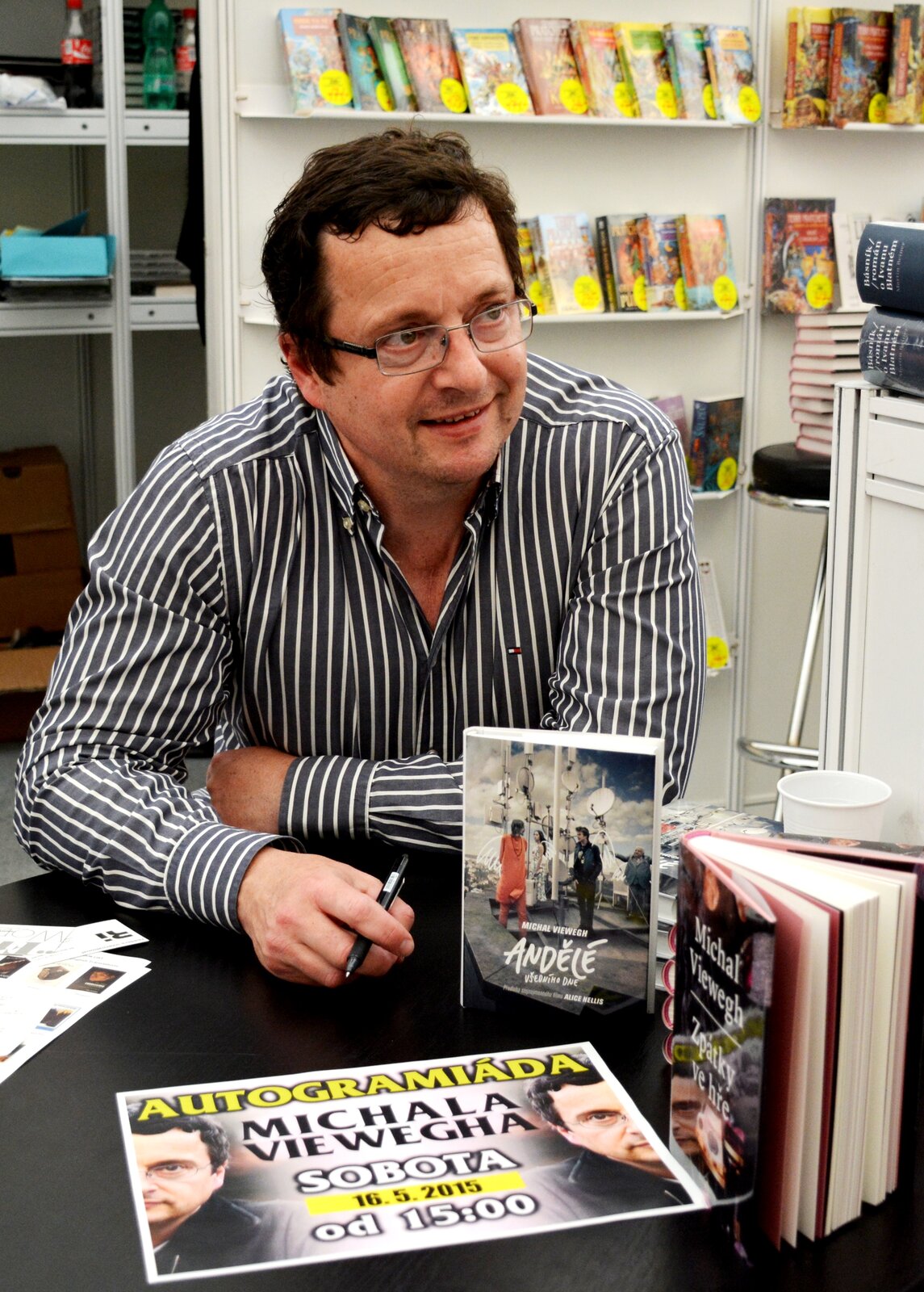 Fotografia barwna w układzie pionowym przedstawiająca w centrum kompozycji czeskiego pisarza Michaela Viewegha siedzącego za stołem. Jest to mężczyzna w średnim wieku. Ma ciemne, trochę lokowane, niezbyt krótko przycięte włosy z dość długimi pejsami i delikatny zarost na okrągłej twarzy. Pisarz jest ubrany w rozchełstaną pod szyją koszulę w czarno‑białe pionowe pasy.