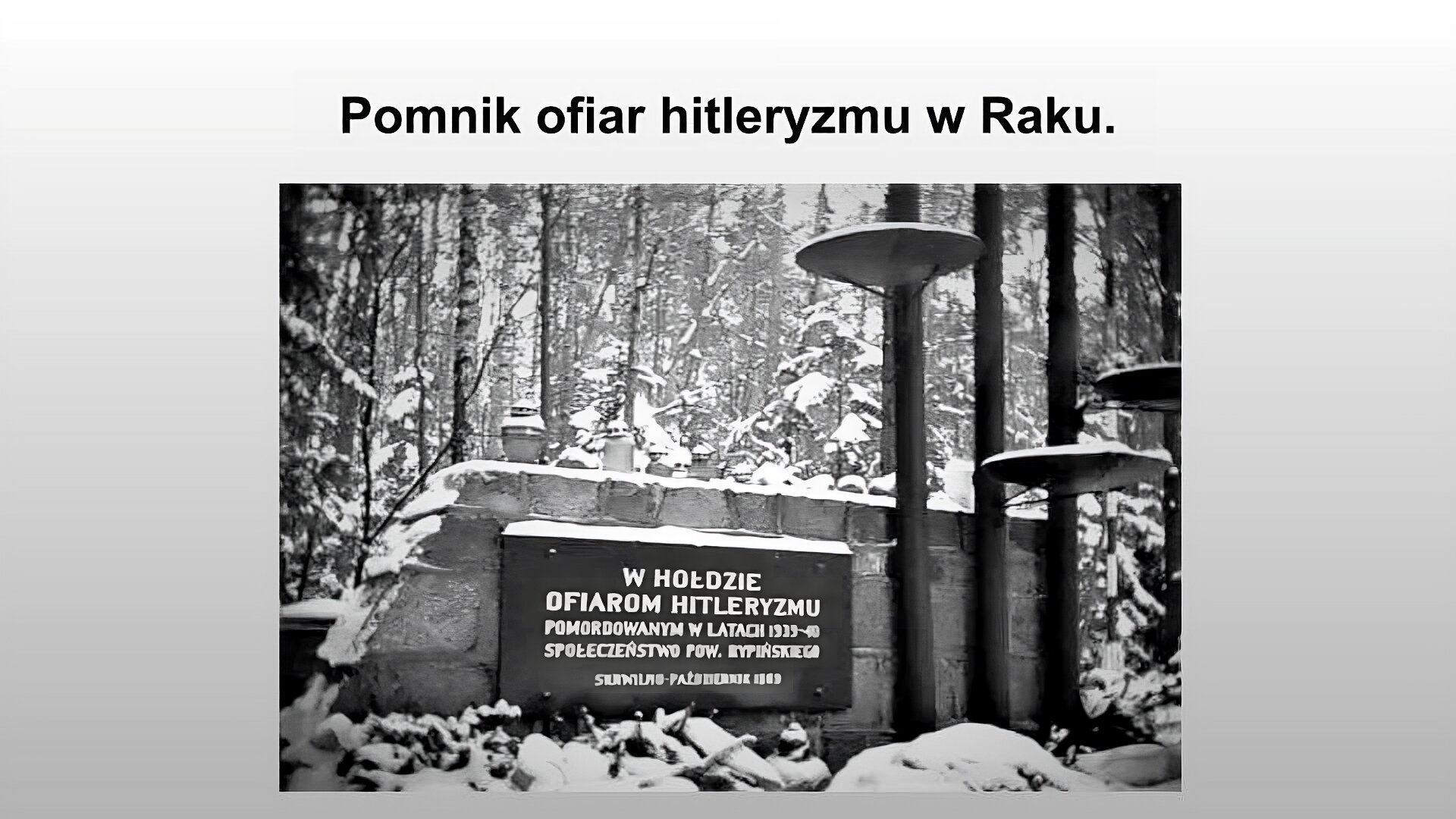 Jasnoszary slajd. Na górze slajdu nagłówek: „Pomnik ofiar hitleryzmu w Raku”. Poniżej czarno- białe zdjęcie przedstawiające kamienny murek. Po prawej stronie trzy ciemne słupy  z zawieszonymi na nich elementami w kształcie talerzy. Na murku czarna tablica z białymi napisami: „W hołdzie ofiarom hitleryzmu pomordowanym w latach 1939–40 społeczeństwo powiatu rypińskiego. Skrwilno – październik 1969”.