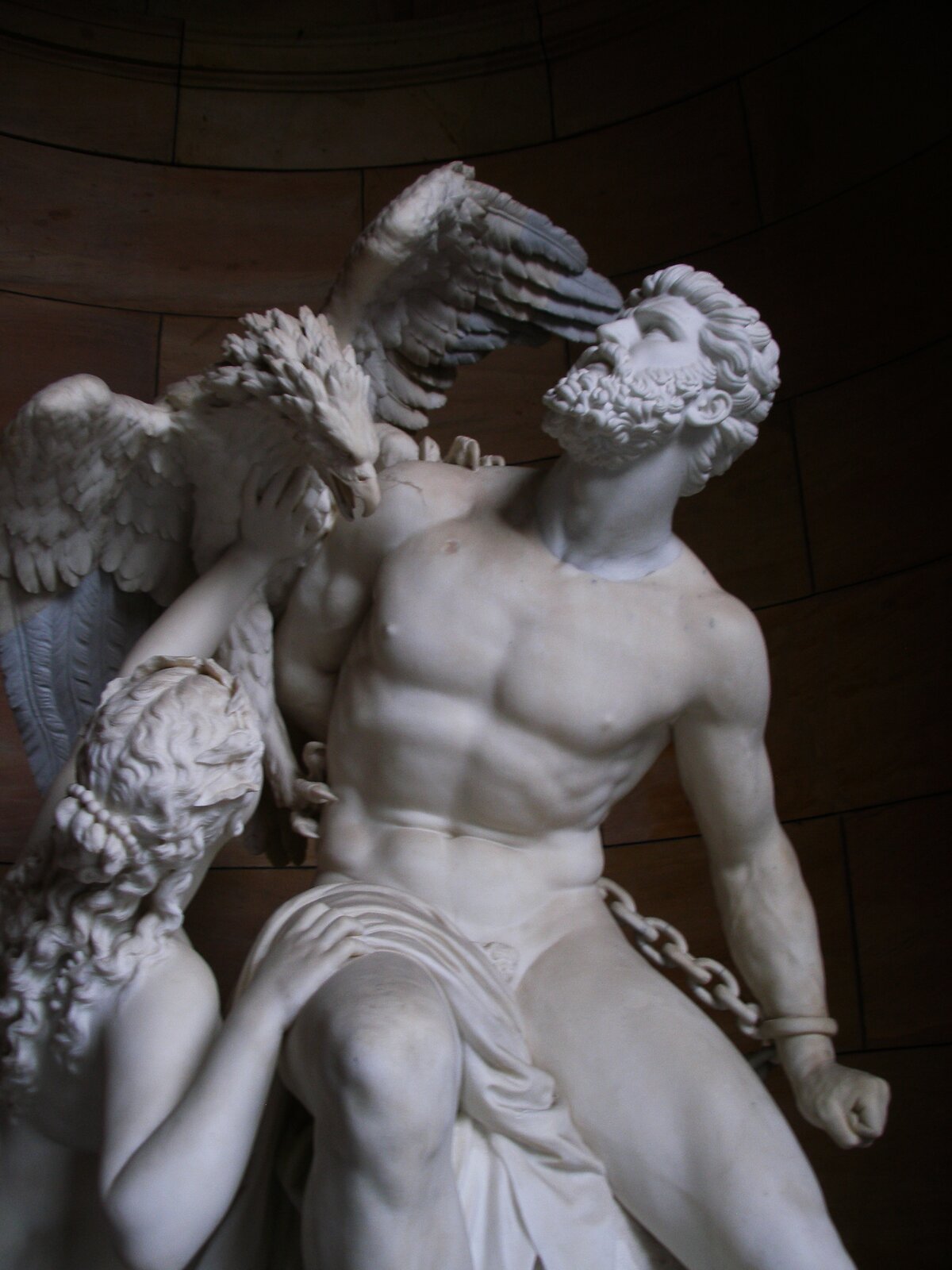 Zdjęcie przedstawia antyczną rzeźbę. To postać siedzącego, atletycznie zbudowanego, nagiego mężczyzny. Ręce ma skute łańcuchem za plecami. U stóp mężczyzny klęczy naga kobieta z długimi włosami. Odpycha od prawego ramienia mężczyzny orła z rozpostartymi skrzydłami. 