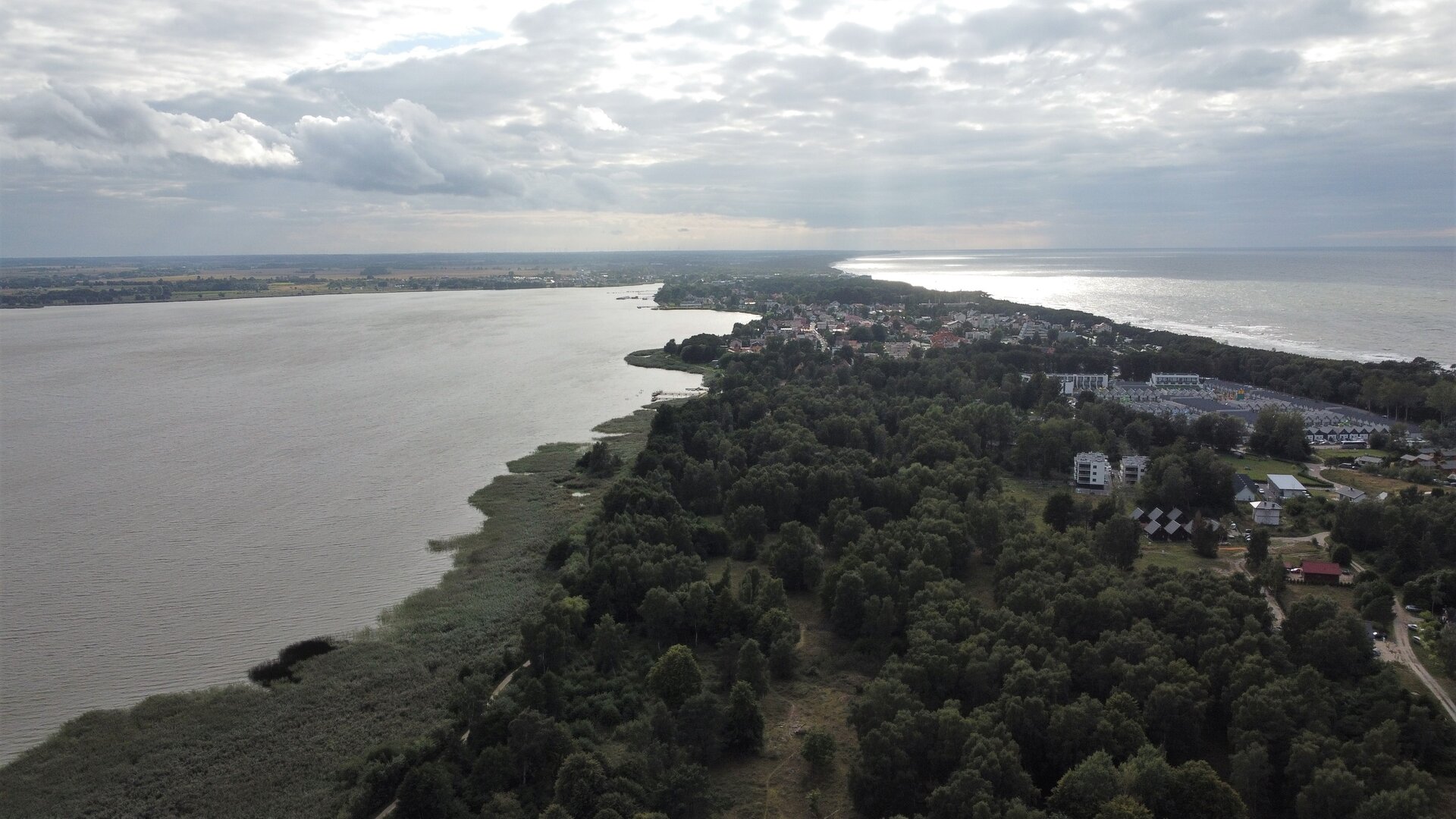 Zdjęcie lotnicze przedstawia po lewej stronie przybrzeżne jezioro, natomiast po prawej stronie rozciąga się morze. Pomiędzy morzem a jeziorem ukazany jest teren porośnięty drzewami, na którym również znajdują się domy.
