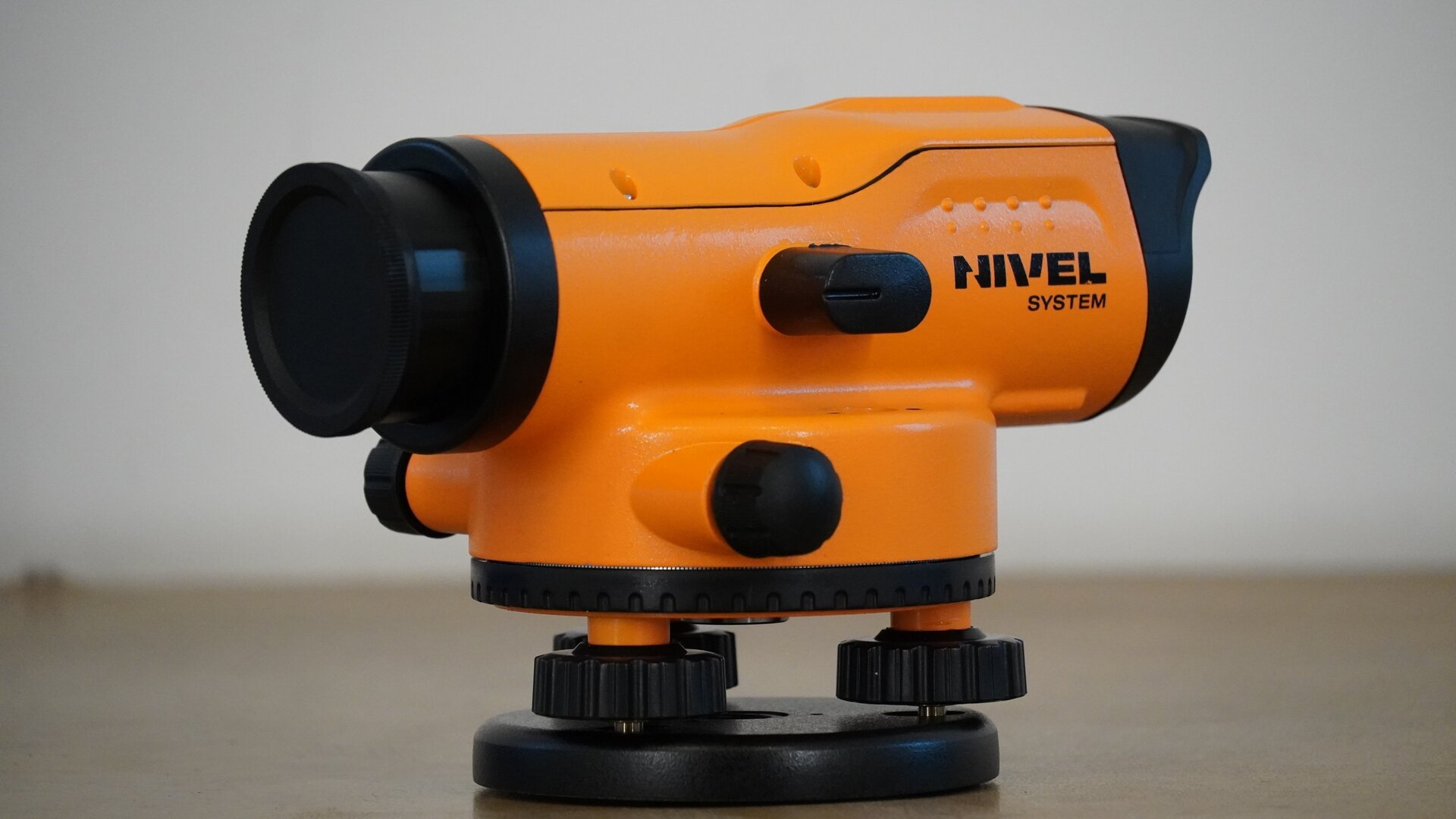 Zdjęcie ukazuje niwelator z innej perspektywy, od strony obiektywu. Na samym dole znajduje się okrągły czarny element, to spodarka. Na niej mieszczą się trzy śruby, na nich zaś koło poziome z podziałką stanowiące dolną część okrągłego korpusu. Z boku korpusu znajduje się czarne pokrętło leniwki, a na korpusie podłużna luneta, która z jednej strony posiada węższy okular, a z drugiej – obiektyw. Z boku lunety mieści się mały owalny element, to libella.