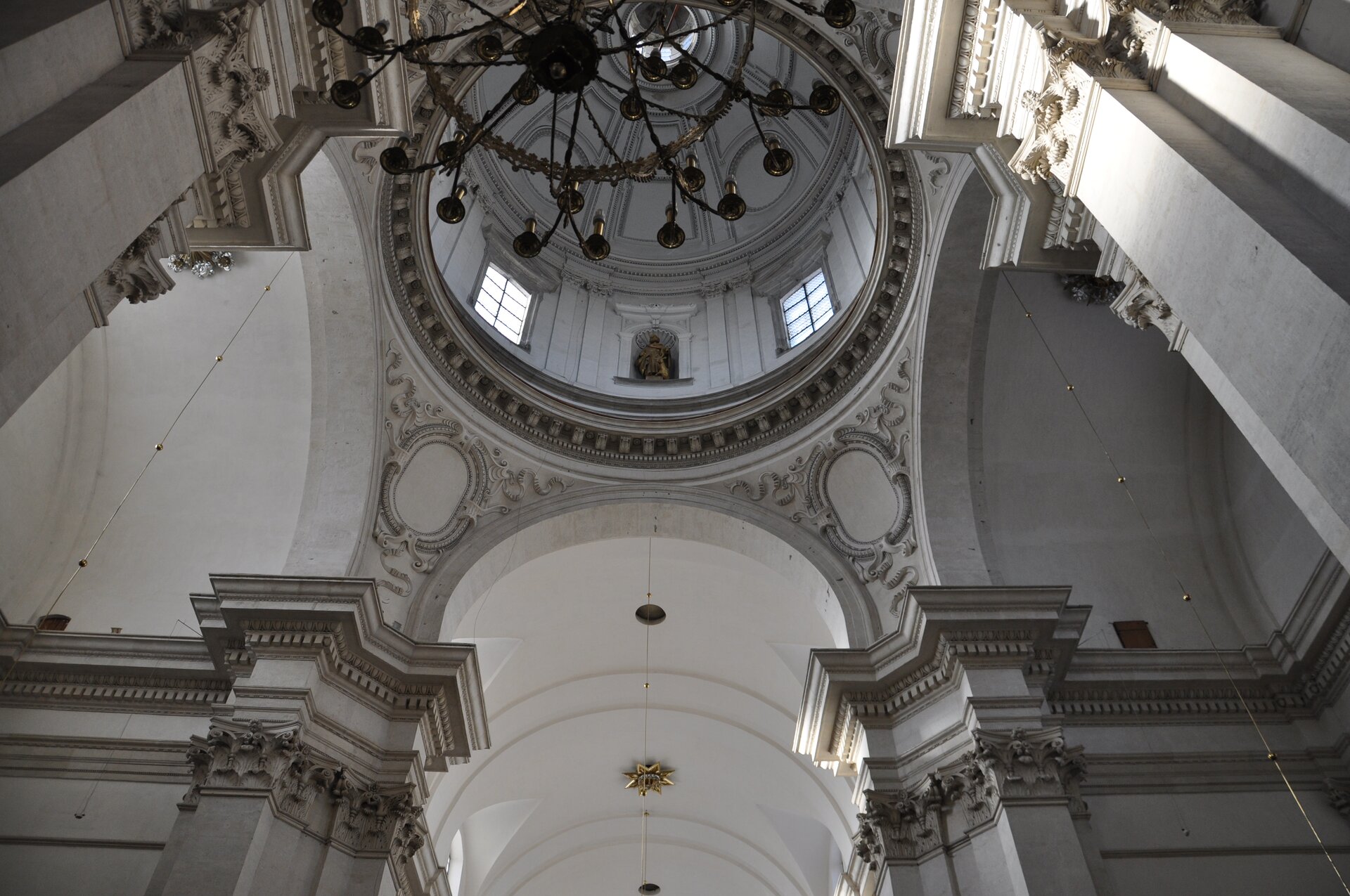 Ilustracja przedstawia kopułę Kościoła pw św. Piotra i Pawła. Na fotografii ukazany jest fragment wnętrza budowli z kopułą oraz górnymi partiami filarów oddzielających nawę od kaplic bocznych. W bębnie kopuły widoczne są okna oraz - naprzemiennie z nimi – nisze z rzeźbami. W kadrze znajduje się również fragment żyrandolu.