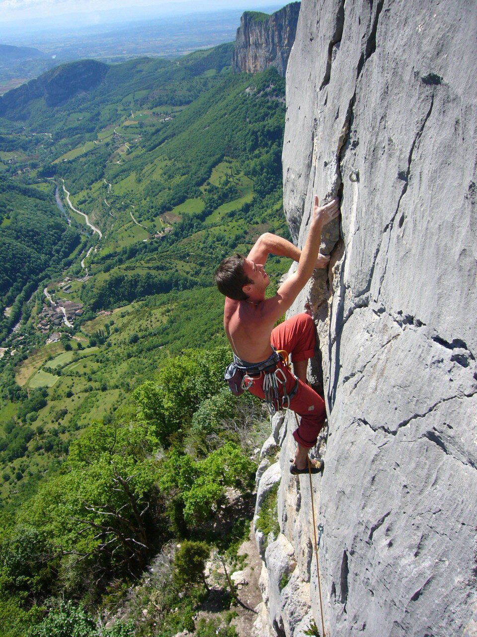 Na zdjęciu znajduje się mężczyzna, który wspina się po skalnej ścianie. Ma nagi tors i czerwone spodnie z przymocowanym do nich sprzętem wspinaczkowym. Po prawej stronie widoczna jest skała, po której się wspina. Lewą część zdjęcia wypełnia widok zielonej doliny w oddali.
