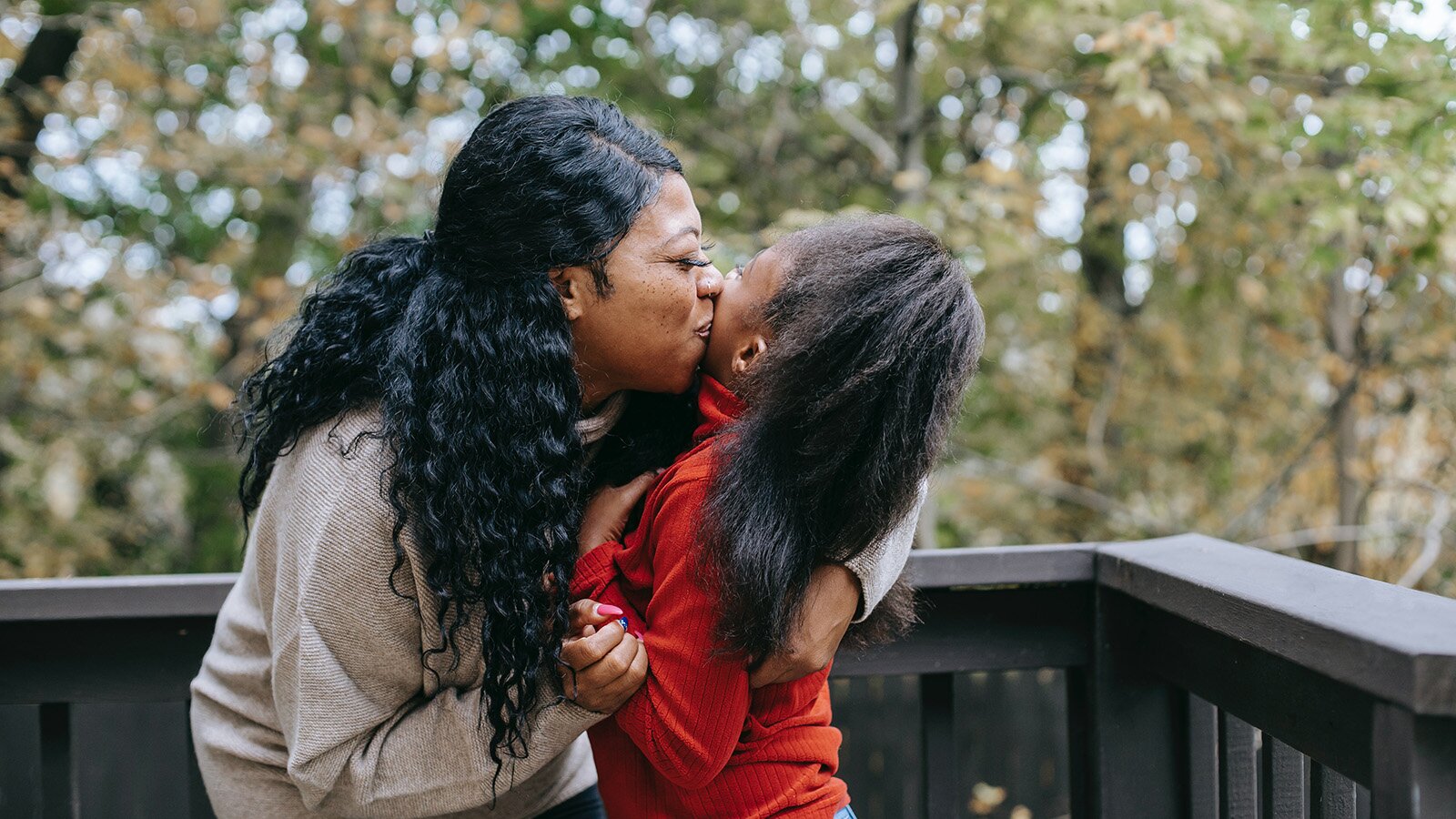 Zdjęcie przedstawia stojącą na tarasie kobietę, która przytula dziewczynkę. Kobieta widoczna po lewej stronie ma długie, pofalowane, czarne włosy i jest ubrana w beżową bluzkę. Kobieta pochyla się i całuje lewy policzek dziecka. Prawą ręką trzyma lewy rękaw bluzki dziewczynki, zaś lewą rękę ma położoną  na jej plecach. Dziewczynka z ciemnymi, długimi włosami, ubrana w czerwoną bluzkę, jest przytulana przez kobietę. W tle widoczne są drzewa.