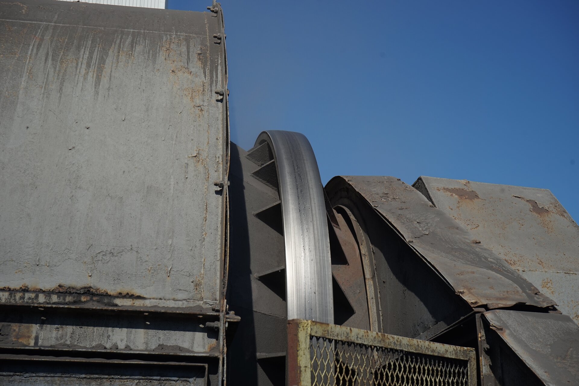 Zdjęcie przedstawia z zewnątrz kruszarkę typu Bradford. Zaprezentowany jest fragment dużego wirującego cylindra. Metalowy pierścień połyskuje. Ziarna w trakcie obrotu bębna kruszarki unoszone są, po czym spadają pod wpływem siły ciężkości, a uderzając o dno. ulegają skruszeniu. Z wnętrza kruszarki wyprowadzona jest na otwarte powietrze skała płonna, niepodlegająca skruszeniu w procesie. Jest ona transportowana przenośnikiem taśmowym znajdującym się na pierwszym planie zdjęcia. Jest on skierowany do przodu. Poniżej zdjęcia widoczny jest przycisk multimedialny z nagraniem audio. Treść nagrania jest zgodna z tytułem zdjęcia.